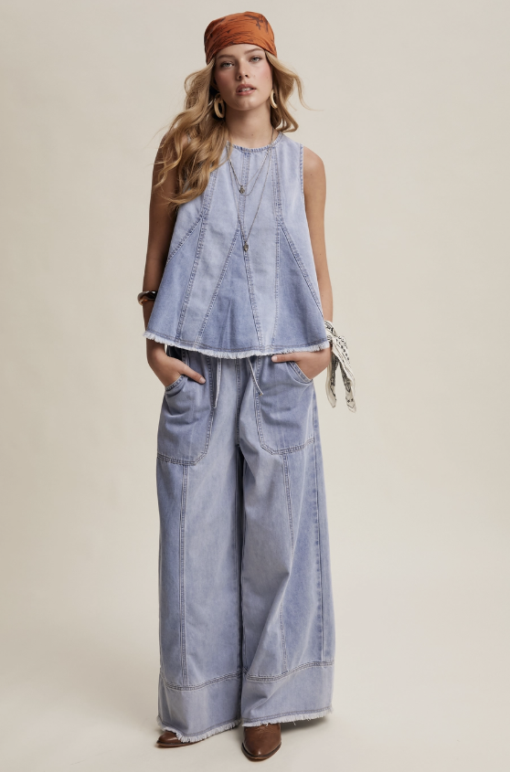 A woman modeling a light blue denim sleeveless top and matching wide-leg pants, styled with an orange-brown headwrap, layered necklaces, hoop earrings, and brown shoes against a plain beige studio background.