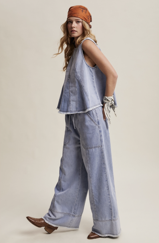 A model wearing a light blue denim vest and matching wide-leg jeans, styled with an orange bandana, white rope bracelet, and brown leather shoes, set against a solid beige studio background.