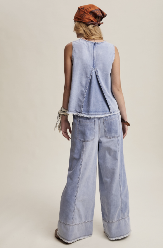A model shown from the back wearing a light blue denim sleeveless vest with a unique stitched back detail and matching wide-leg denim pants, paired with an orange-brown patterned headscarf, against a solid beige studio background.