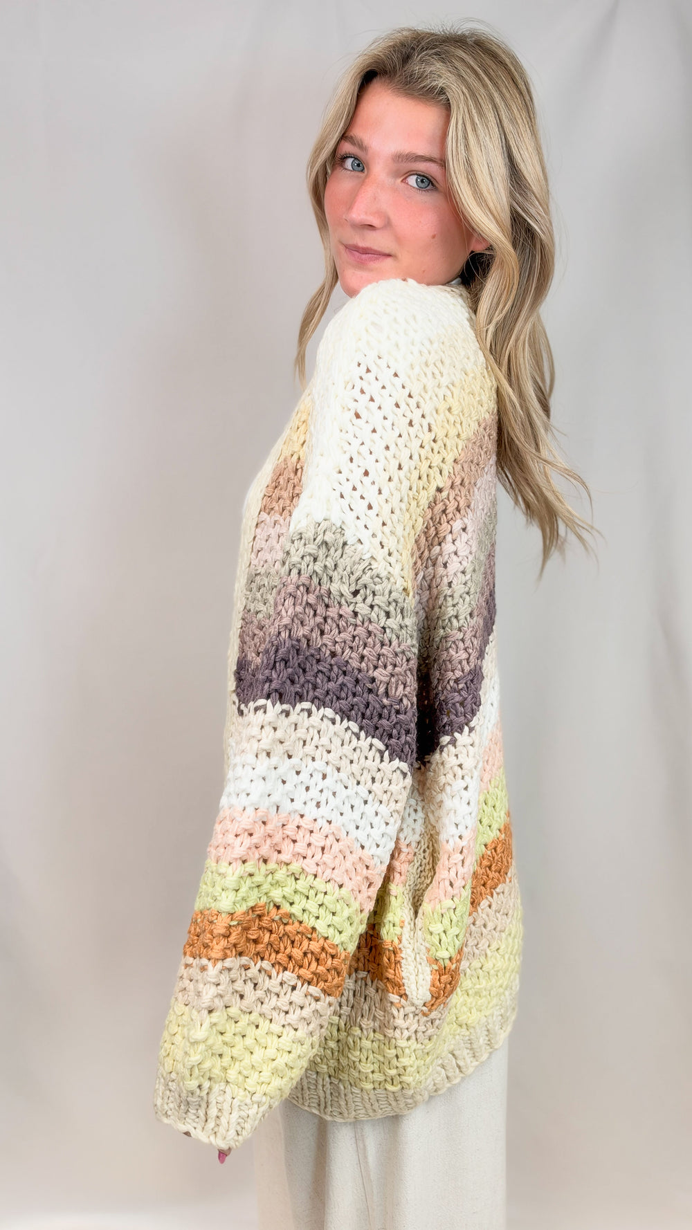Woman shown from side view wearing chunky knit sweater with multi color stripes on plain abckground