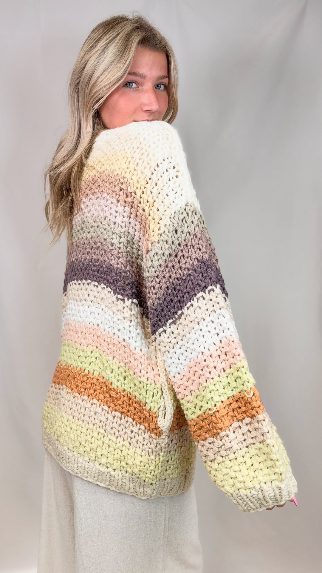 Woman shown from side view wearing chunky knit cardigan with multi color stripes on plain background