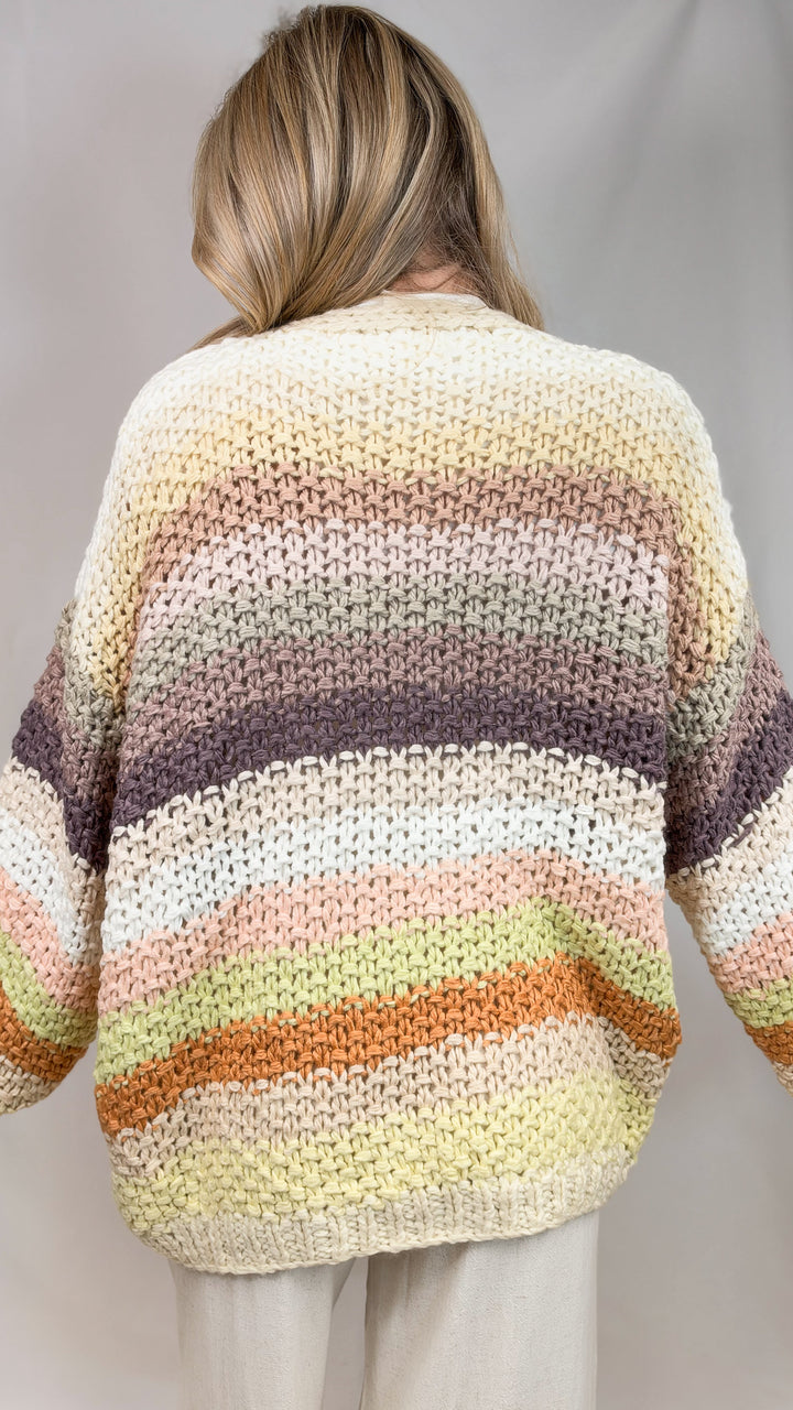 Woman shown from back view wearing chunky knit sweater with multi color stripes on plain background
