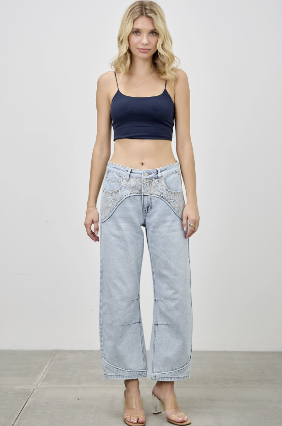 A woman modeling a pair of light-wash wide-leg cropped jeans with decorative contrast stitching and a navy blue crop top, standing against a plain white studio wall.