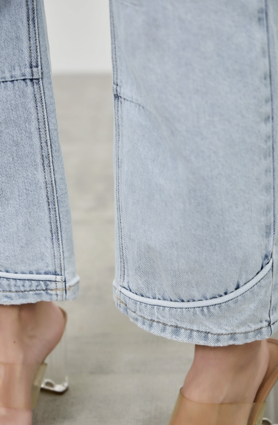 Close-up of a person wearing light-wash wide-leg denim jeans with rolled hems and clear heeled sandals, shown against a neutral indoor floor background.