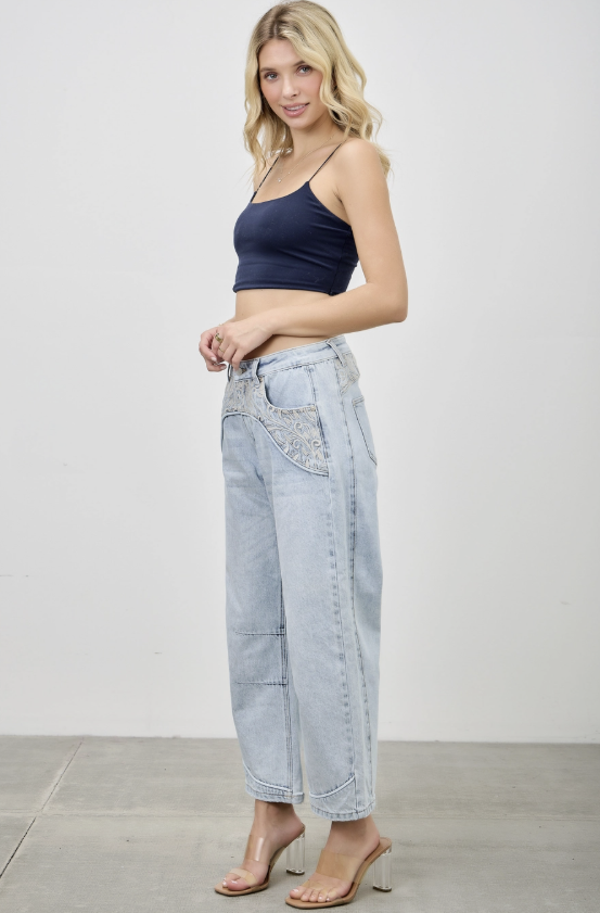 A woman modeling light-wash wide-leg denim jeans with decorative embroidered details on the front pockets, paired with a navy blue crop top and clear high-heeled sandals, set against a plain white studio background.