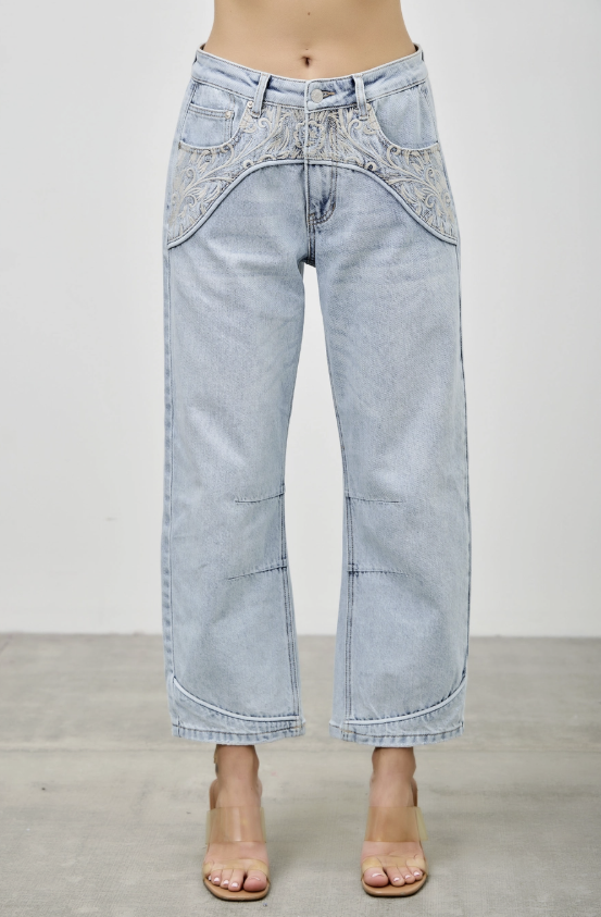 A model is wearing a pair of light-wash blue denim jeans with unique patchwork paneling and embroidery on the front, paired with clear high-heeled sandals against a neutral studio background.
