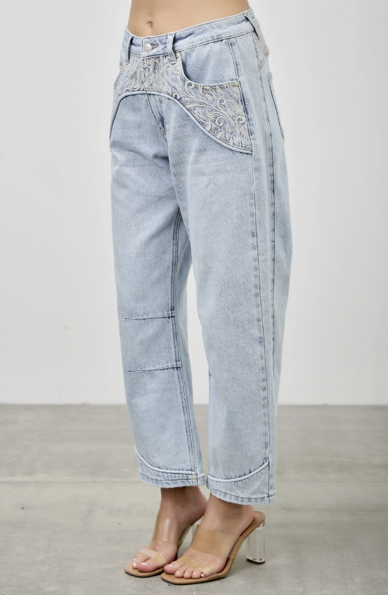 Light wash blue denim cropped pants with decorative embroidered detailing across the front pockets, shown on a model wearing clear-strap heeled sandals against a neutral studio background.