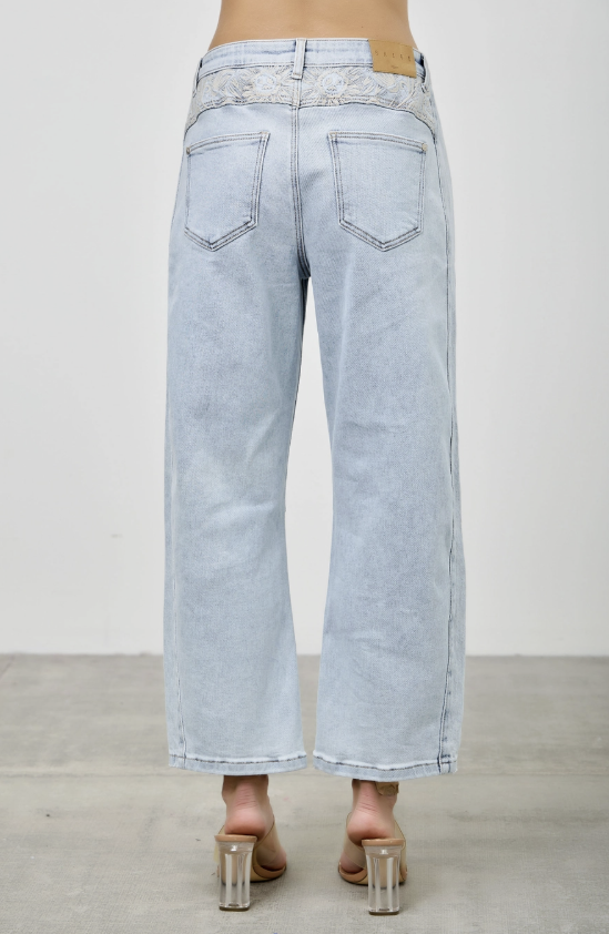Back view of a person wearing light-wash wide-leg cropped jeans with a lace panel detail at the back waistband, paired with clear-strap heeled sandals against a plain indoor wall and concrete floor.