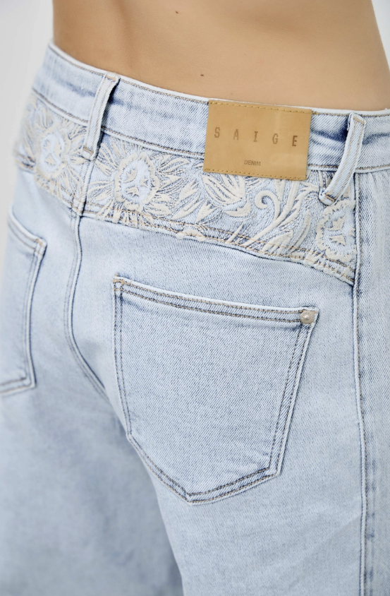 Close-up of the back waistband of light-wash blue jeans with white embroidered detailing and a tan leather patch reading SAIGE.