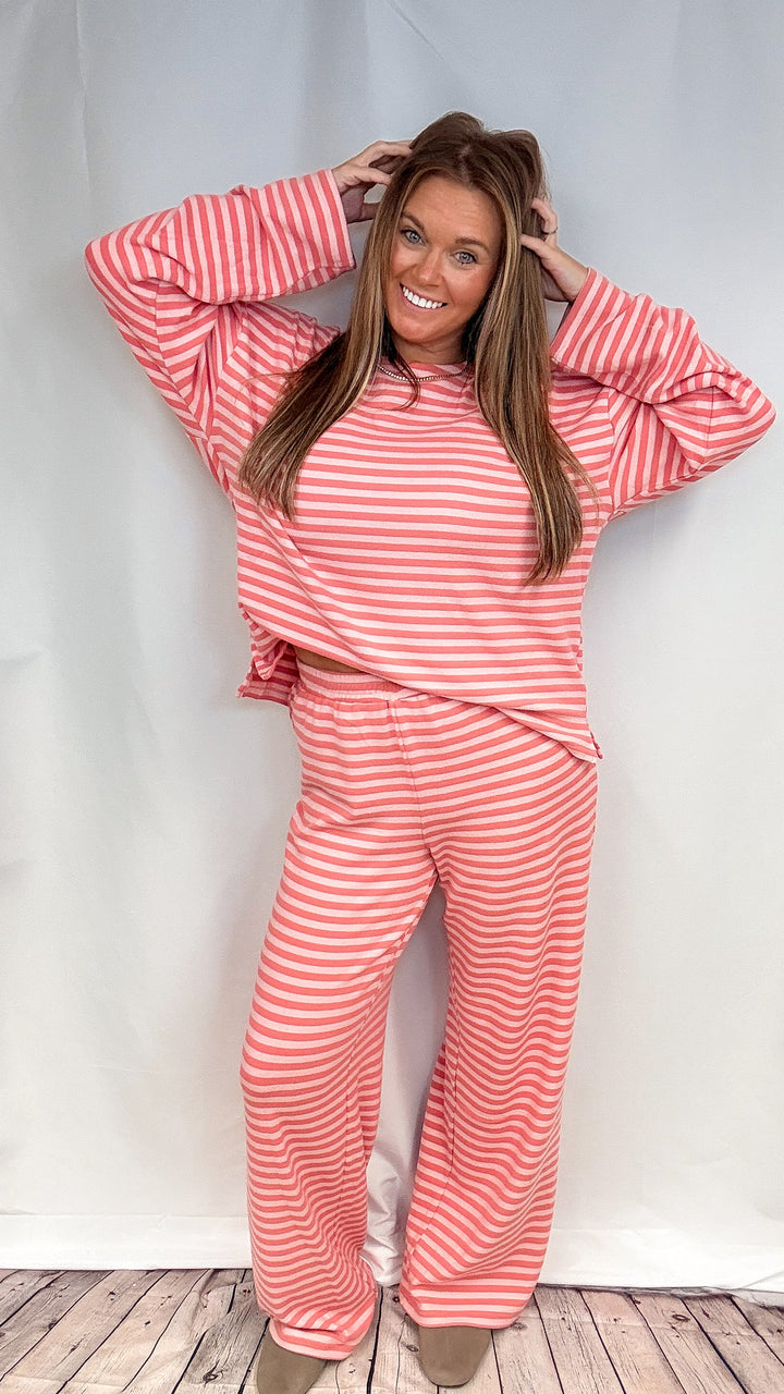 A woman modeling a matching two-piece loungewear set with pink and white horizontal stripes, consisting of a long-sleeve cropped top and wide-leg pants, posed against a white studio backdrop.