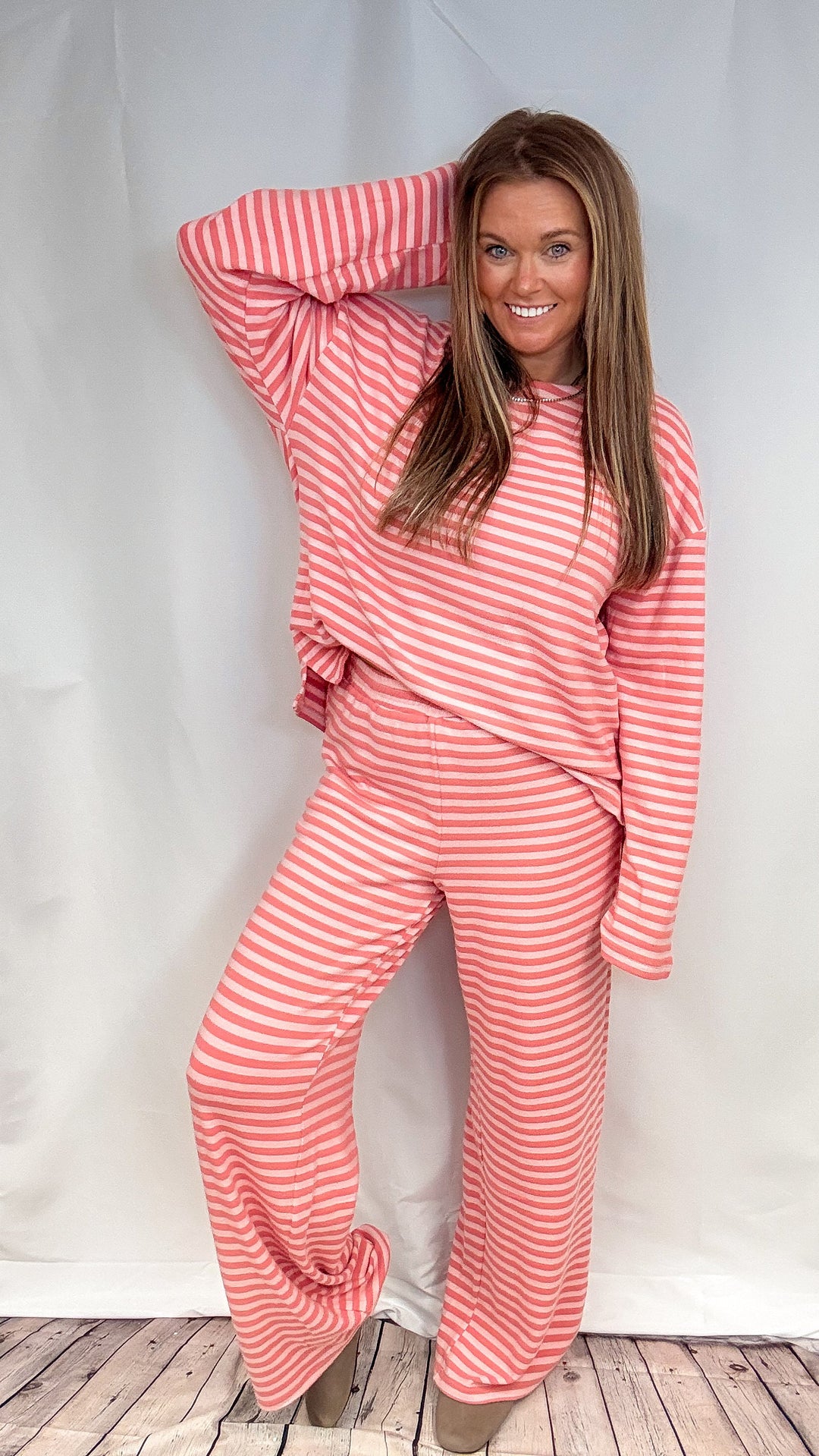 A woman modeling a matching coral pink and white striped lounge set, consisting of a long-sleeve top and wide-leg pants, posed against a white fabric backdrop.