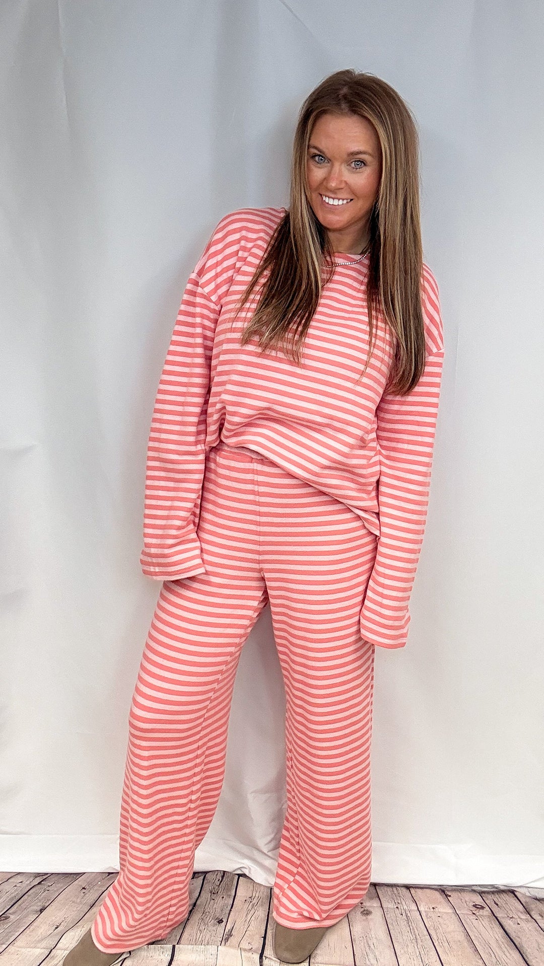 A woman modeling a matching two-piece loungewear set consisting of a long-sleeve top and wide-leg pants, both featuring a pink and white horizontal stripe pattern, set against a white studio backdrop.