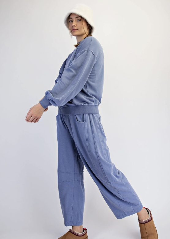 A model wearing a matching dusty blue lounge set consisting of a long-sleeve sweatshirt and wide-leg pants, styled with a white knit hat and brown slip-on slippers against a plain white studio background.