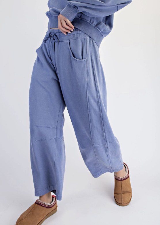 A person wearing loose, wide-leg blue sweatpants with a drawstring waist and brown suede slip-on slippers, photographed against a clean light gray studio background.