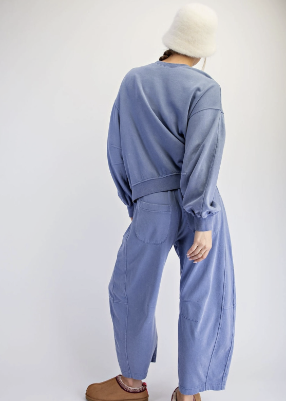 A person shown from the back wearing a dusty blue cropped sweatshirt and matching wide-leg pants, with a white fuzzy hat and brown slippers, against a plain light gray studio background.