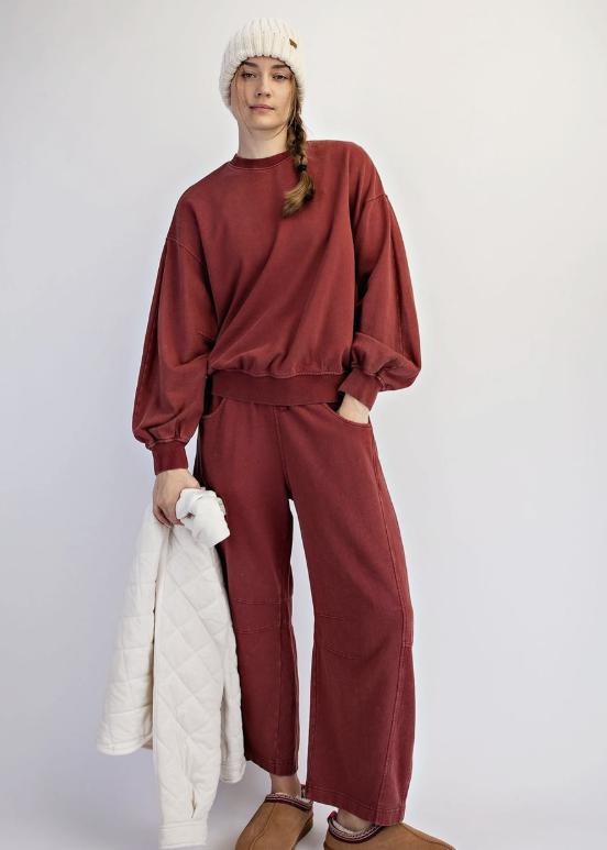 A model stands against a light gray studio background wearing a matching rust-red sweatshirt and wide-leg pants set, a cream knit beanie, brown slippers, and holding a cream quilted jacket.