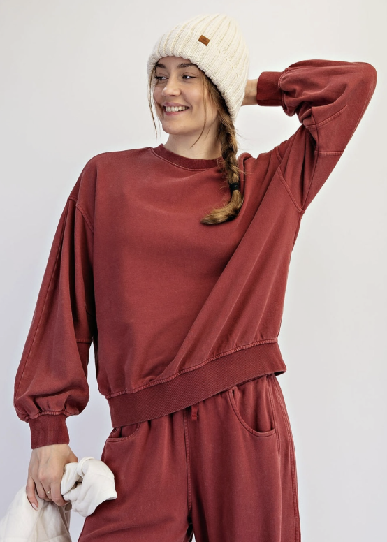 A woman modeling a matching rust-red sweatshirt and sweatpants set, styled with a cream beanie and holding a white quilted jacket against a plain light gray background.