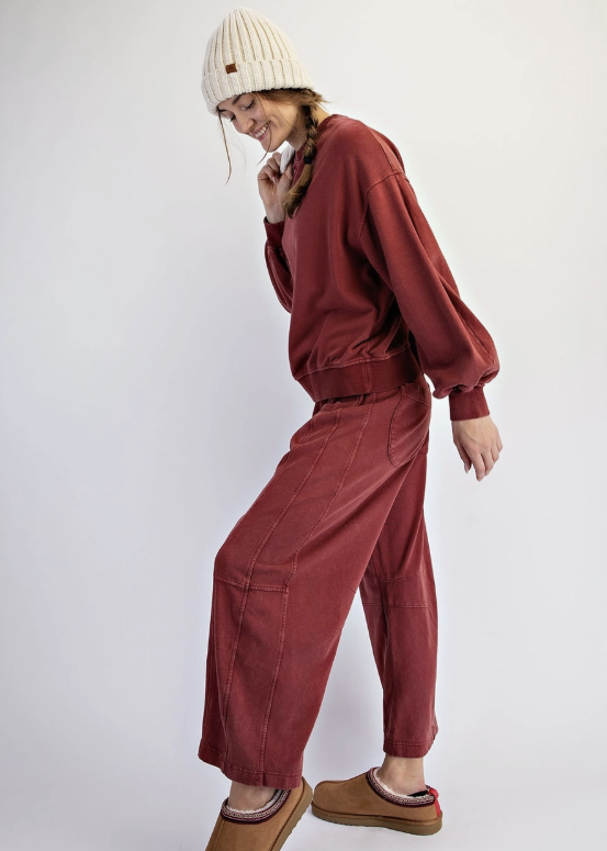A model wearing a matching rust-red sweatshirt and wide-leg pants set, styled with a cream ribbed beanie and brown slippers, posing against a light gray studio background.