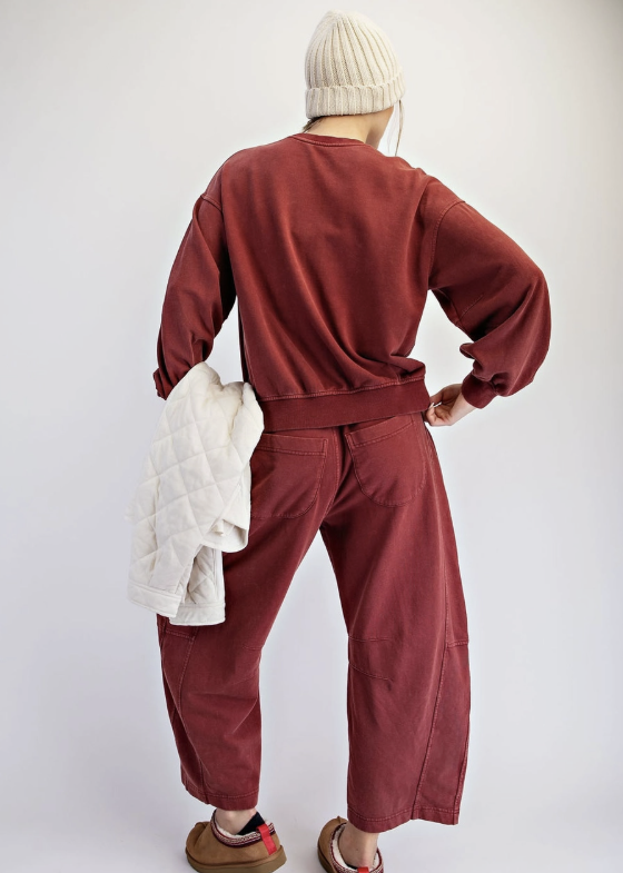 A person shown from the back wearing a matching rust-red sweatshirt and wide-leg pants set, with a cream knit beanie, brown slip-on shoes, and a white quilted jacket draped over one arm, against a plain light gray studio background.