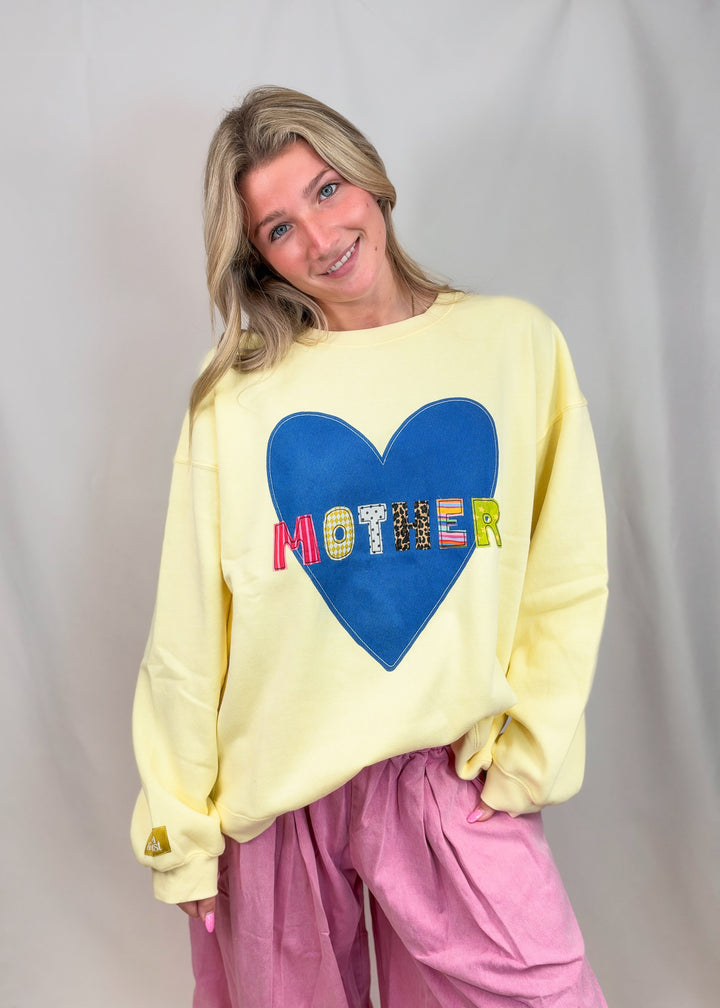 A woman wearing a pale yellow oversized sweatshirt with a large blue heart on the front and the word &