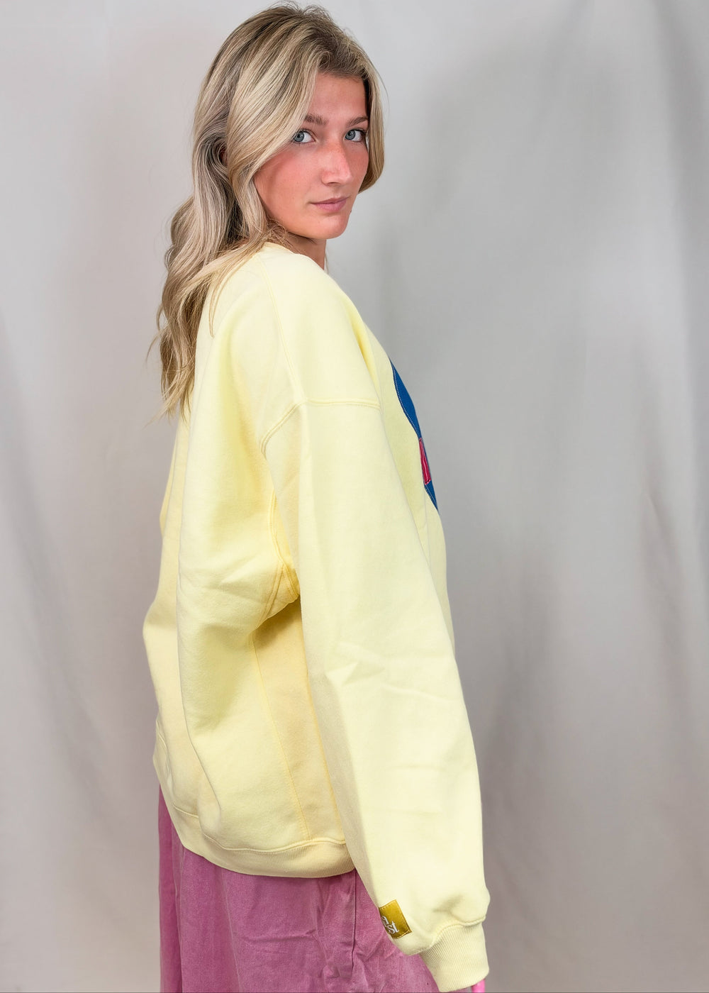 A woman wearing a pale yellow oversized sweatshirt and a pink skirt, photographed against a light gray fabric backdrop.