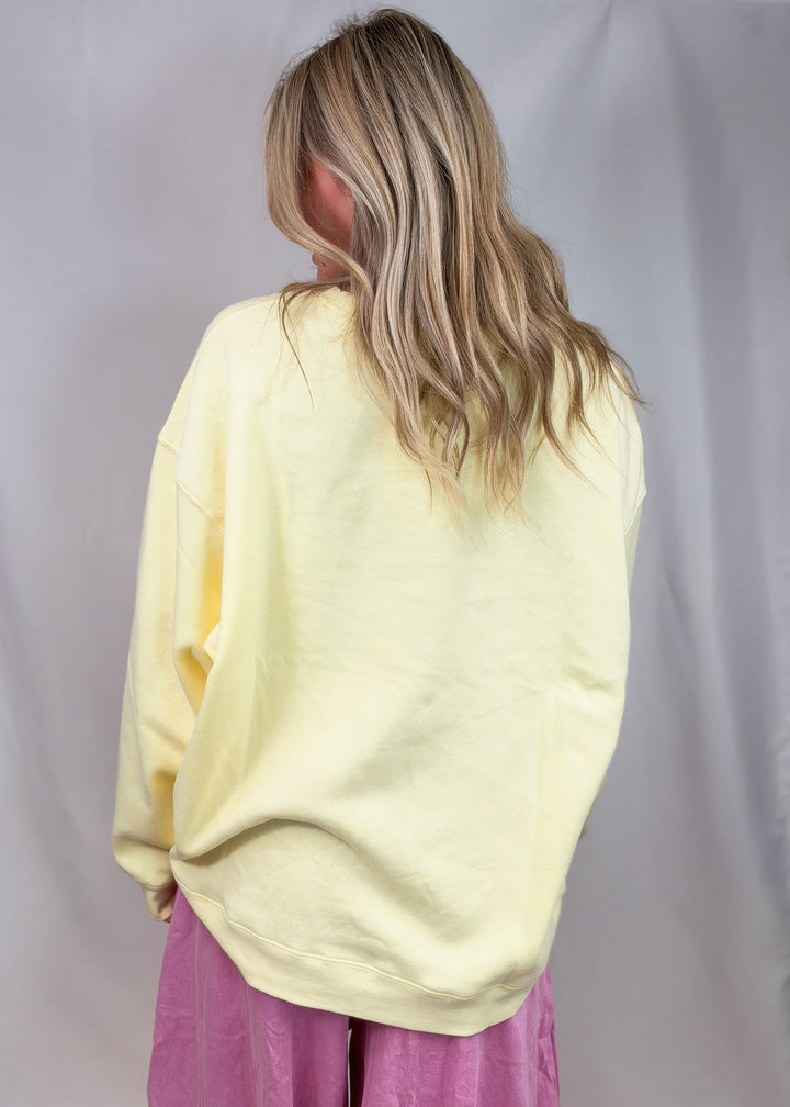 A woman with blonde hair is shown from behind wearing an oversized pale yellow sweatshirt and pink pants, standing against a light gray fabric backdrop.