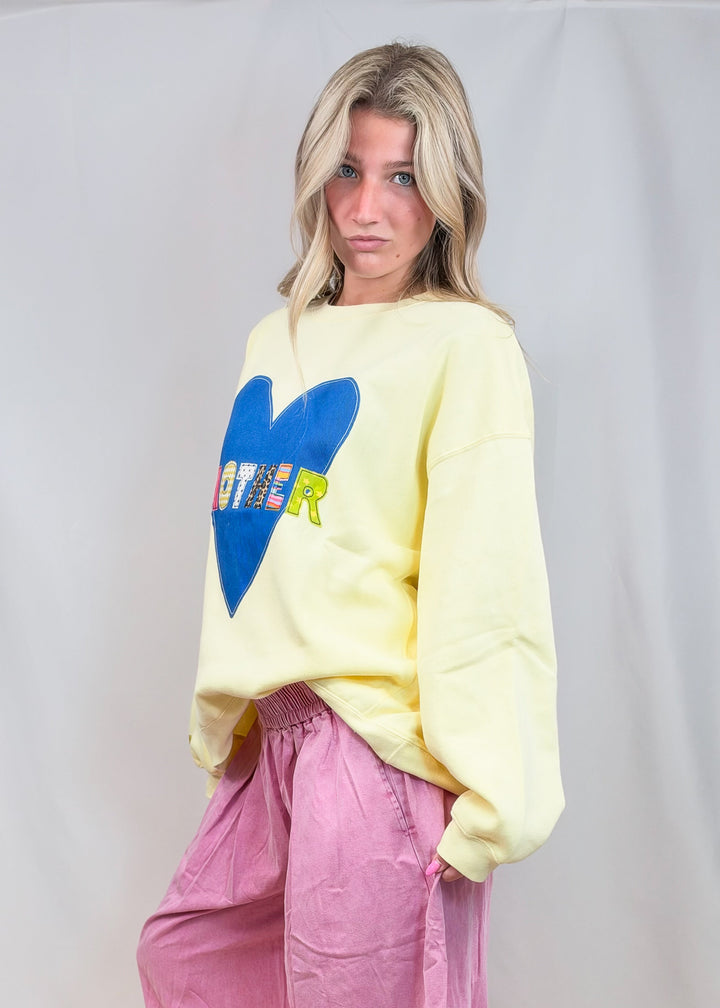 A woman stands barefoot against a light gray studio backdrop, wearing an oversized pale yellow sweatshirt with a large blue heart graphic and the word &