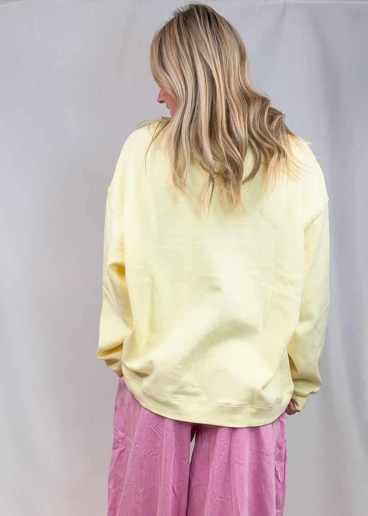 A woman shown from behind wearing a pale yellow oversized sweatshirt and loose pink harem-style pants, standing against a plain light gray studio backdrop.