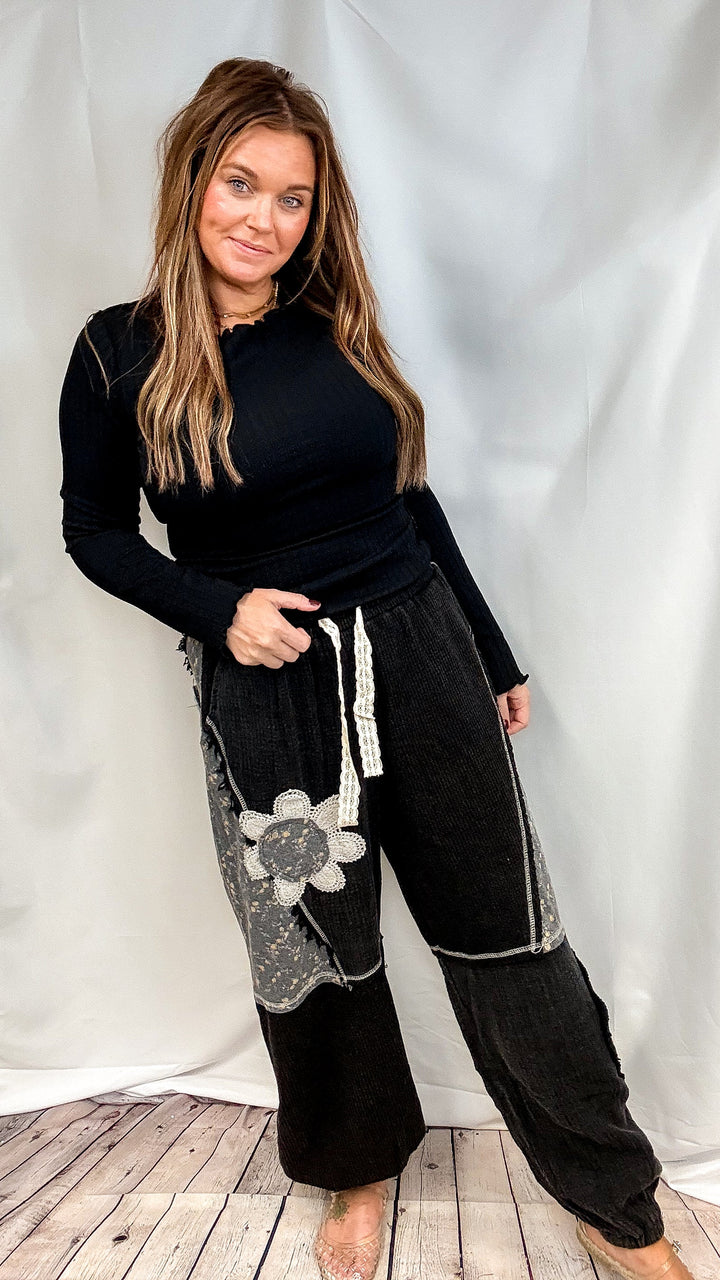 Woman wearing black long sleeve ribbed top with black pants featuring floral patchwork on plain background