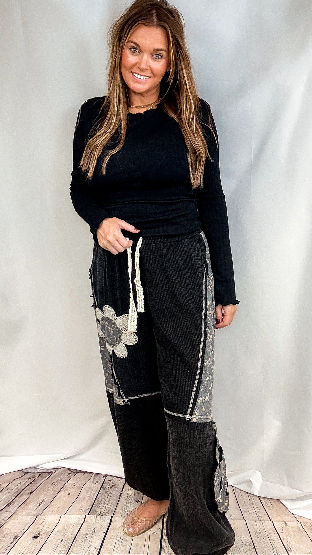 Woman wearing black long sleeve ribbed top and black pants with floral patchwork on plain background