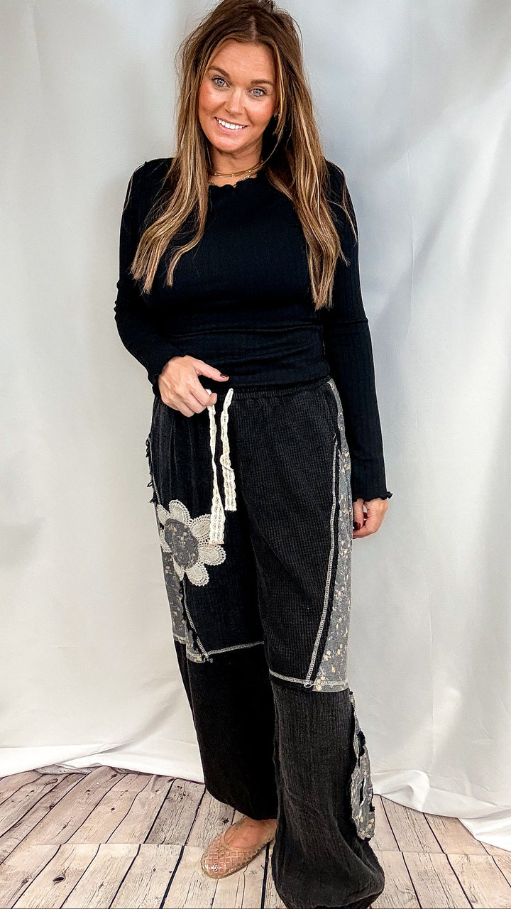 Woman wearing black long sleeve ribbed top and black pants with floral patchwork on plain background