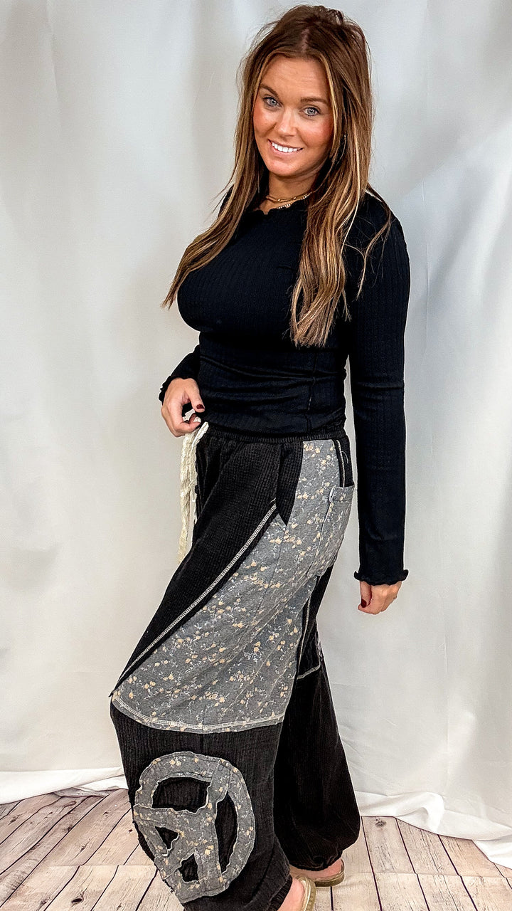 Woman wearing black long sleeve ribbed top and black pants with floral patchwork on plain background