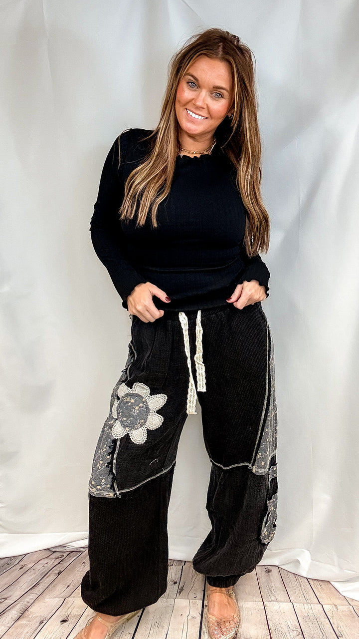 Woman wearing black long sleeve ribbed top and black pants with floral patchwork on plain background