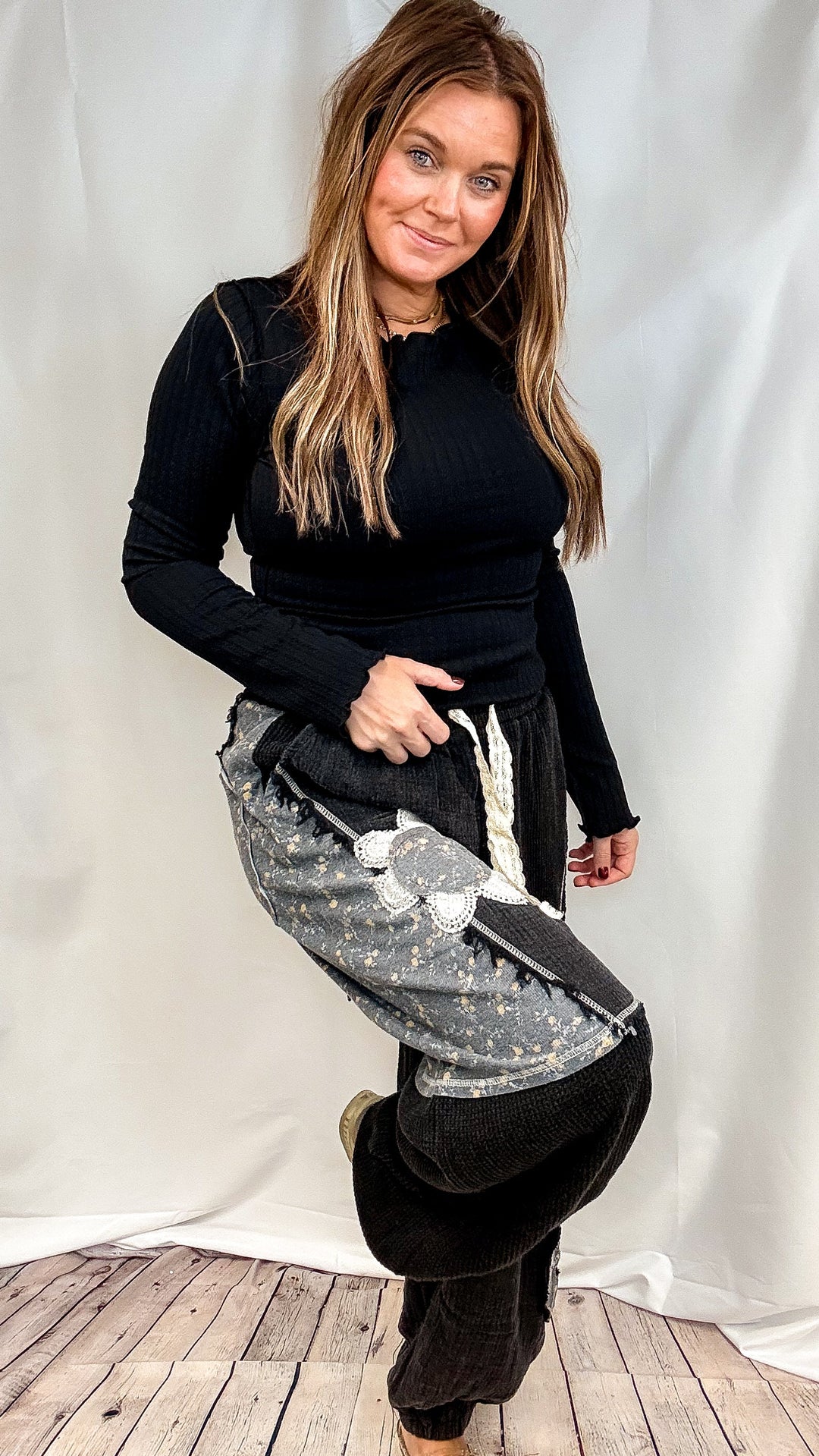 A woman modeling a black ribbed long-sleeve top and gray patterned wide-leg pants with a white graphic print, standing against a white draped backdrop.