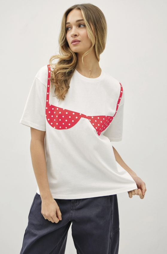 A woman wearing a white short-sleeve t-shirt with a red polka-dot bra graphic on the front, paired with dark blue pants, against a light gray studio background.