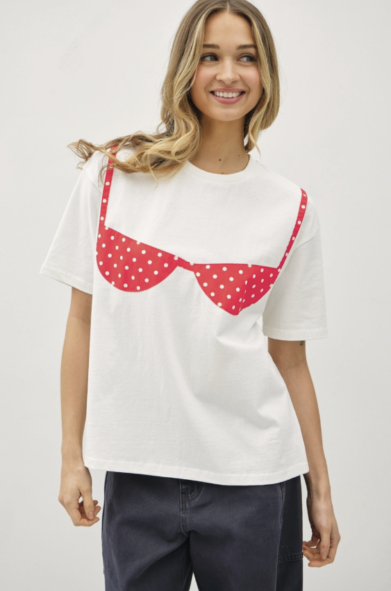 A woman wearing a white short-sleeve t-shirt with a red and white polka dot bra graphic on the front, paired with dark blue pants, against a light gray studio background.