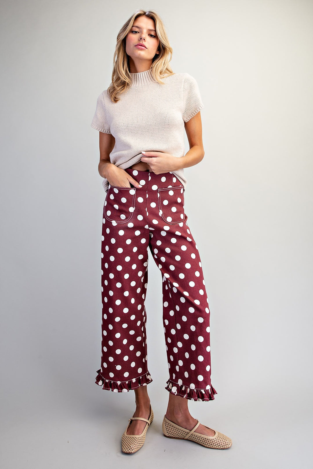 A woman modeling a pair of burgundy wide-leg pants with white polka dots, paired with a cream short-sleeve knit top and beige woven flats, against a light gray studio background.