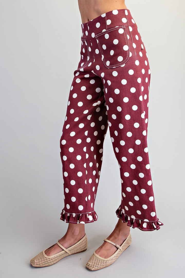 A person wearing burgundy pants with a white polka dot pattern, ruffled hems, and beige woven flat shoes against a light gray studio background.