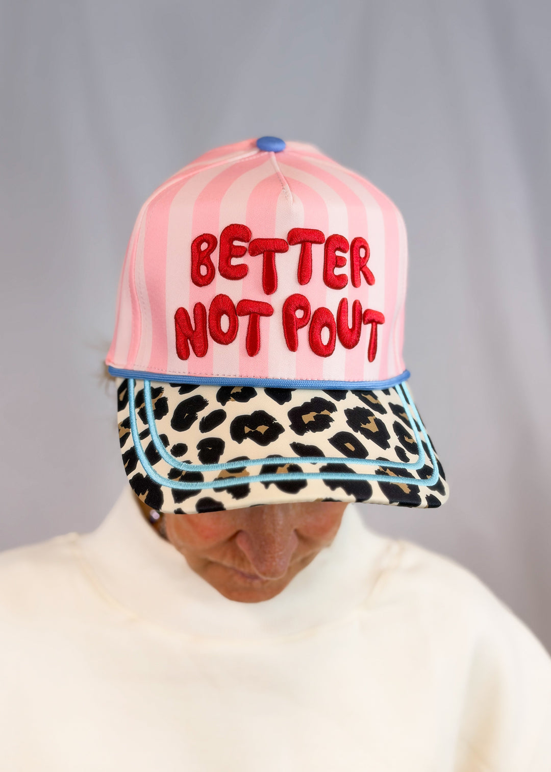 Woman wearing pink stripe trucker hat with 'BETTER NOT POUT' embroidered across front with cheetah bill on plain background
