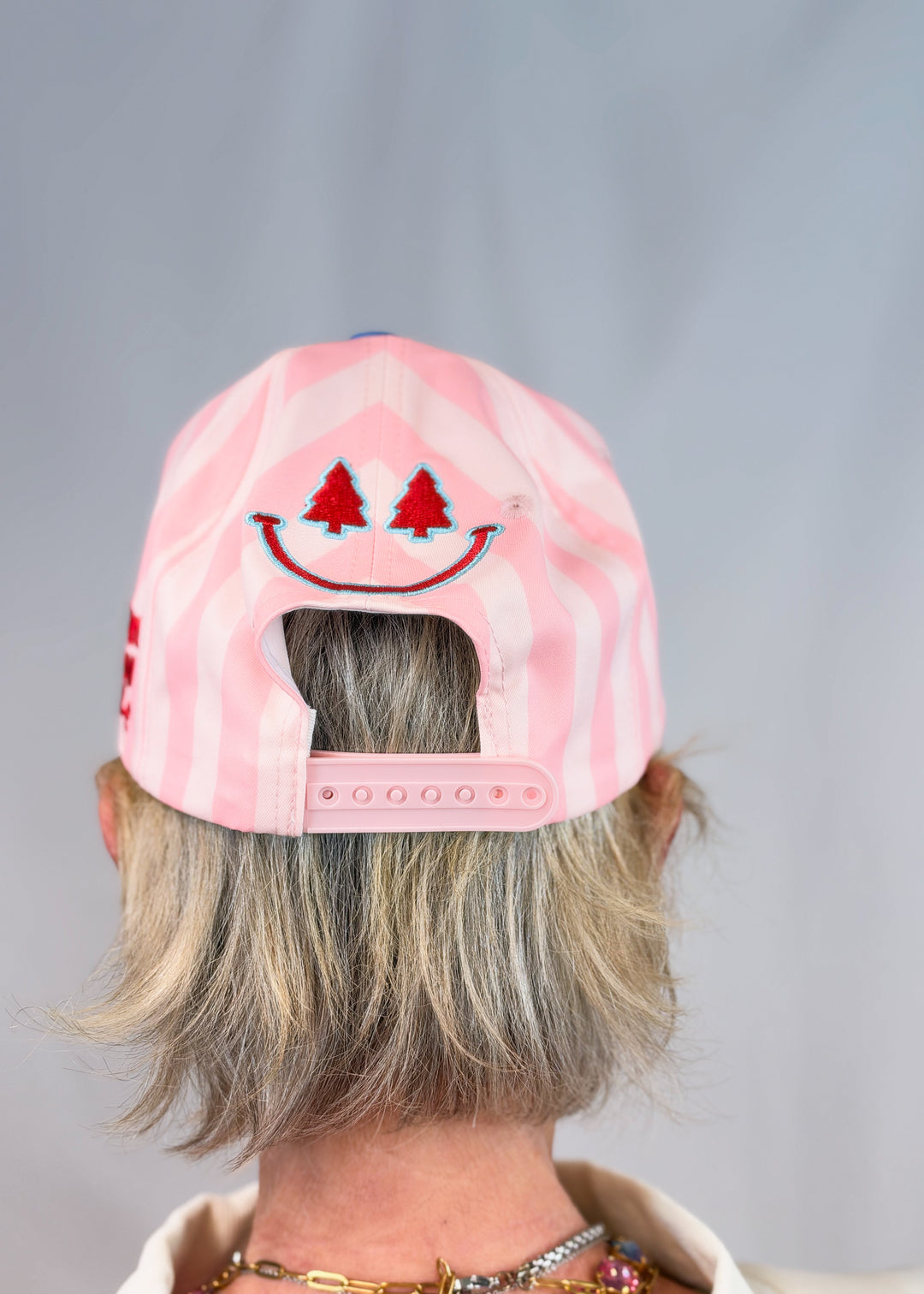 Woman shown from back wearing pink stripe trucker hat with smiley face embroidered on back on plain background