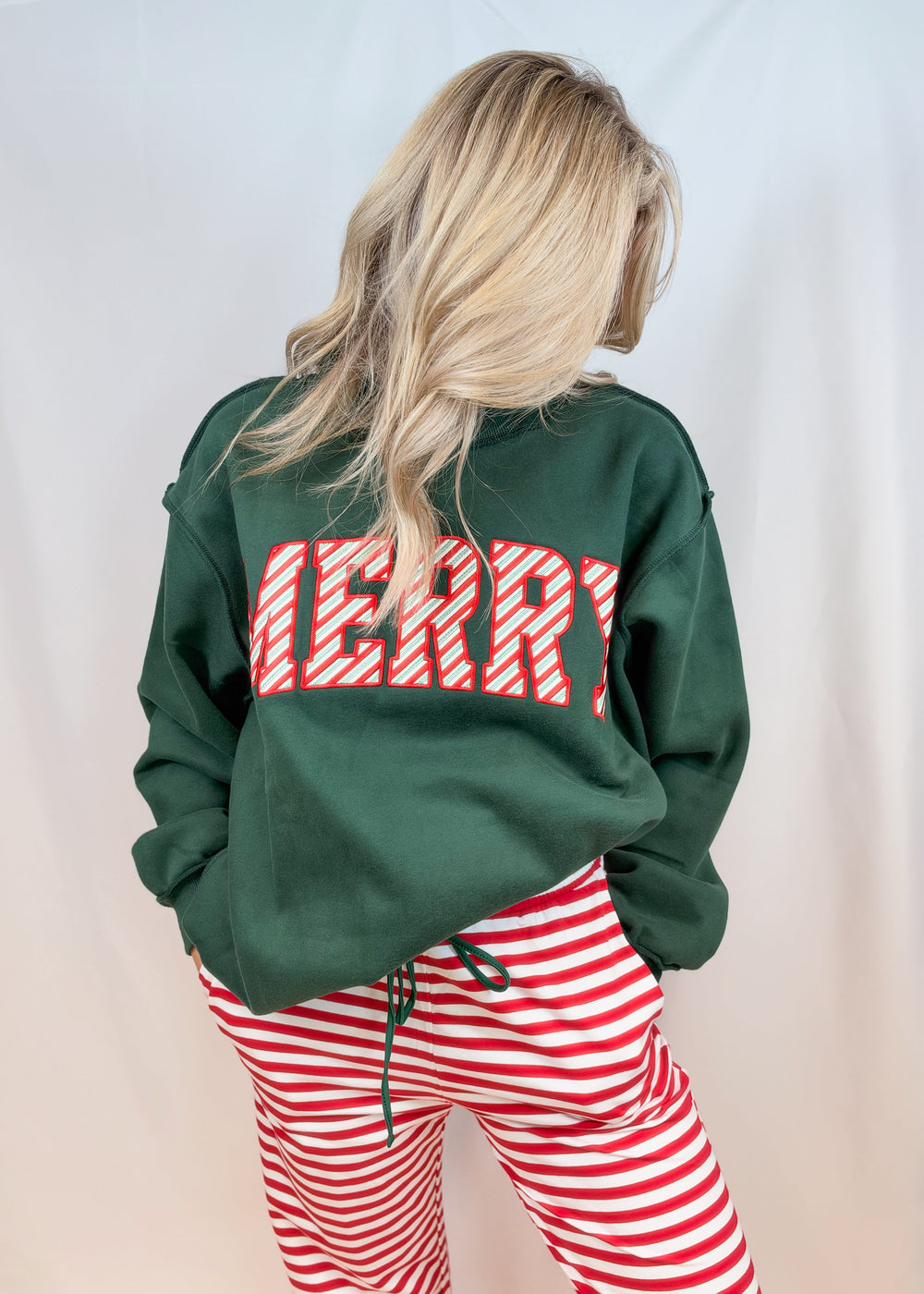 Woman wearing green mock neck sweatshirt with red and white stripe 'MERRY' on front with red and white stripe pants on plain background