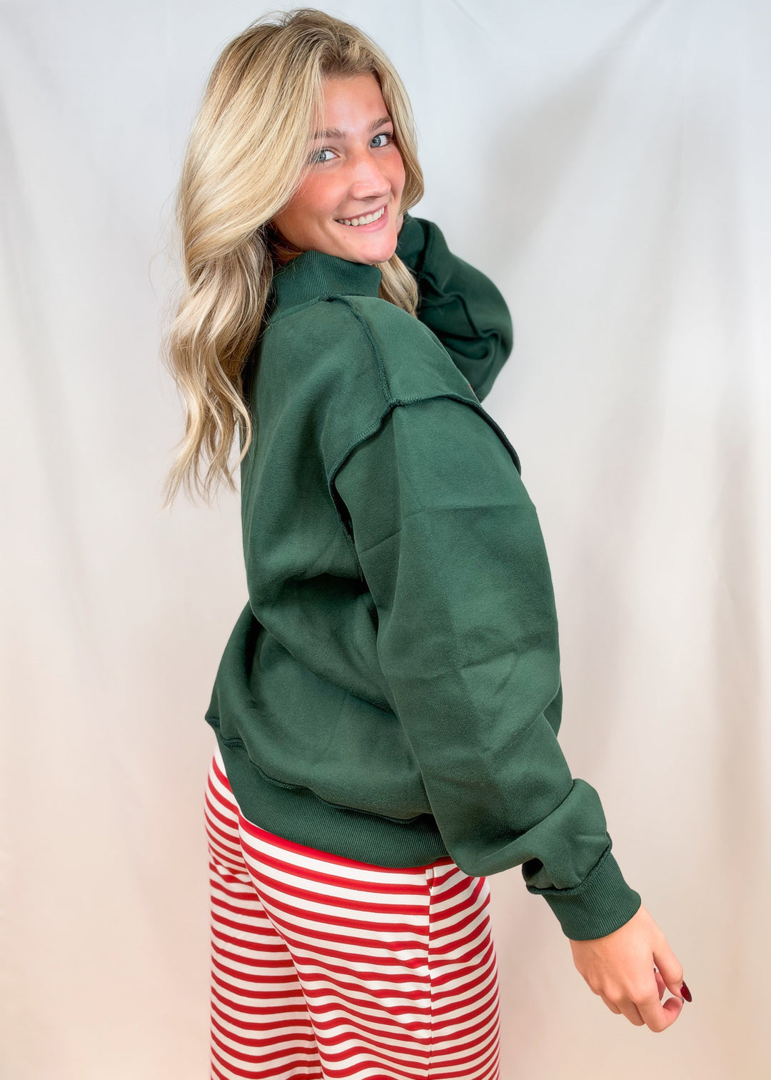 Woman shown from side view wearing green mock neck sweatshirt and red and white stripe pants on plain background