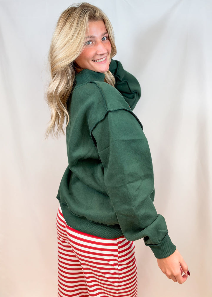 Woman shown from side view wearing green mock neck sweatshirt and red and white stripe pants on plain background