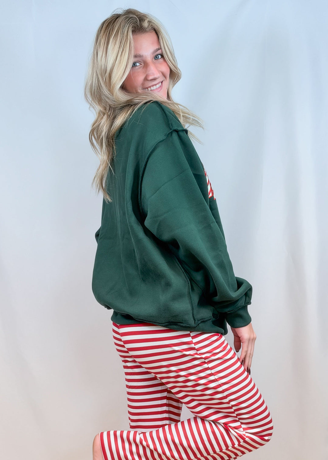 Woman shown from side view wearing green mock neck sweatshirt with red and white stripe pants on plain background