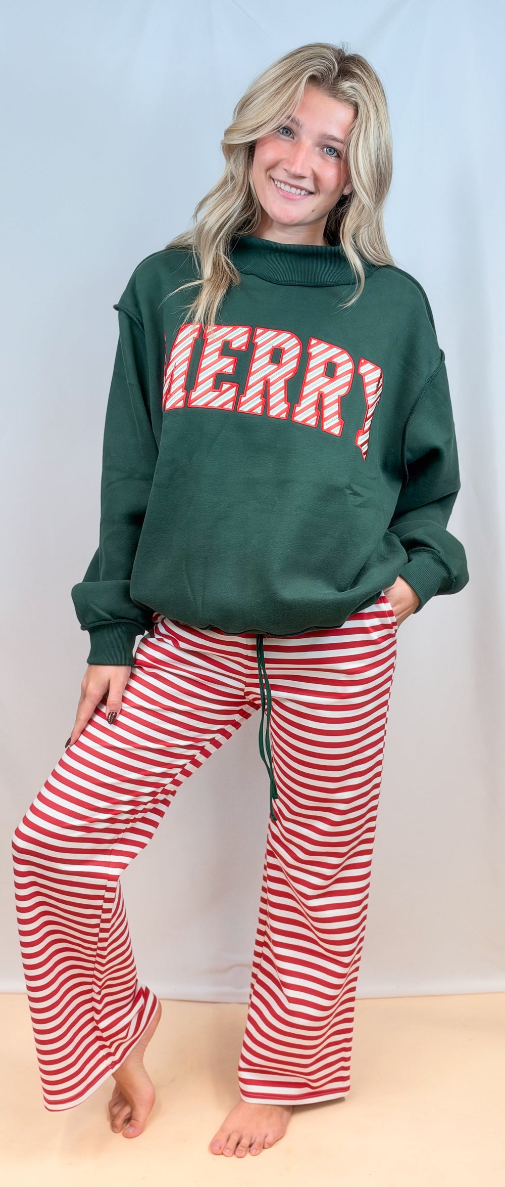 Woman wearing green mock neck sweatshirt with 'MERRY' on front with red and white stripe pants on plain background