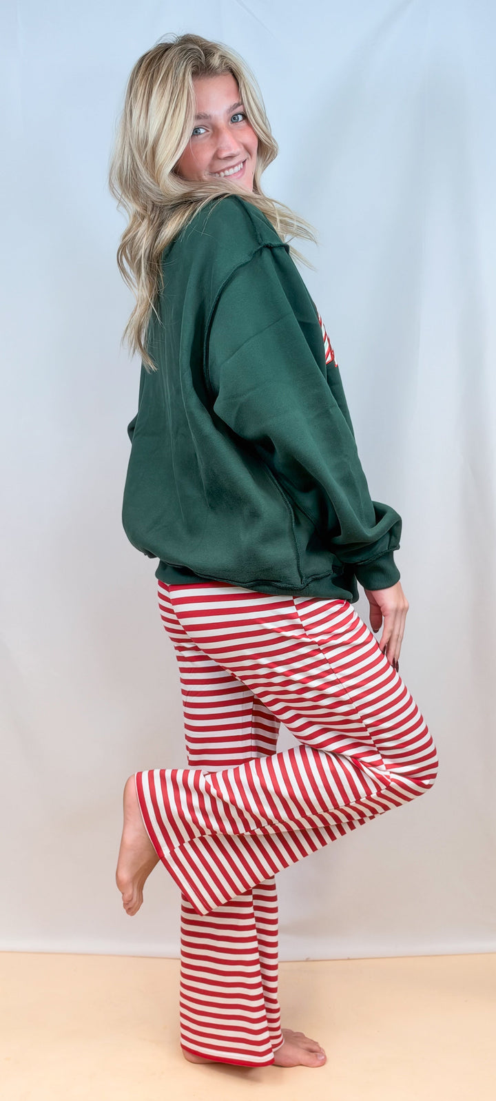 Woman shown from side view wearing green sweatshirt with red and white stripe pants on plain background