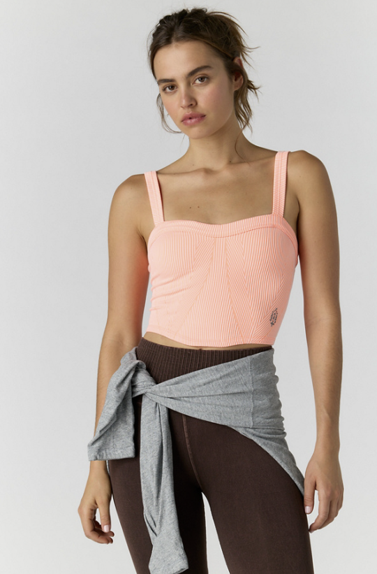 A woman modeling a ribbed scoop-neck tank top in light peach pink, paired with dark brown leggings and a heather gray shirt tied around her waist, against a light gray studio background.