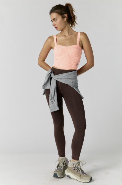 A woman modeling activewear consisting of a light pink ribbed tank top, dark brown leggings, and a light gray sweatshirt tied around her waist, paired with beige hiking boots against a light gray studio background.