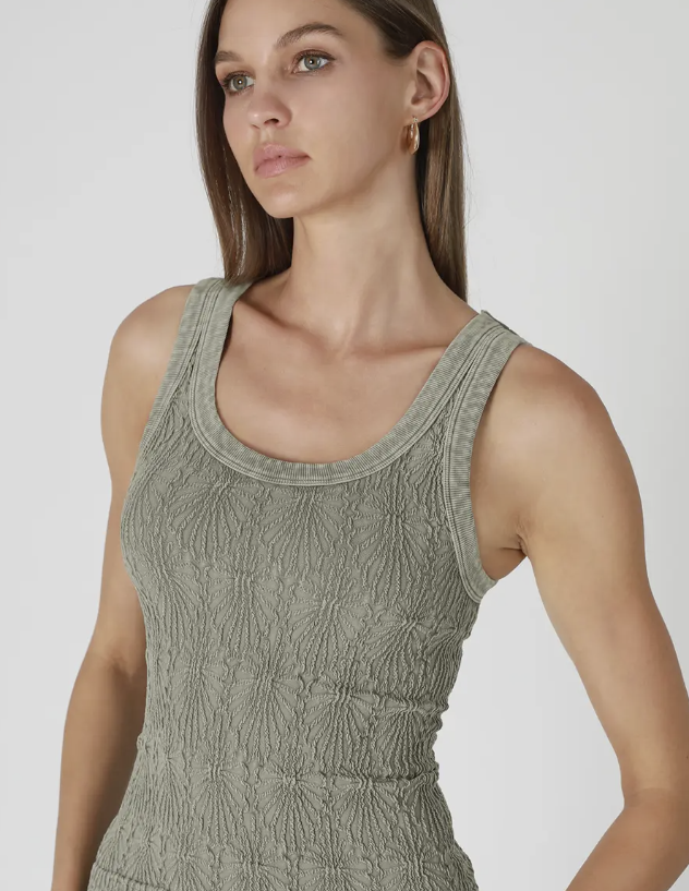 A close-up of a woman wearing a textured, ribbed knit scoop-neck tank top in a muted sage green color, set against a plain light gray studio background.