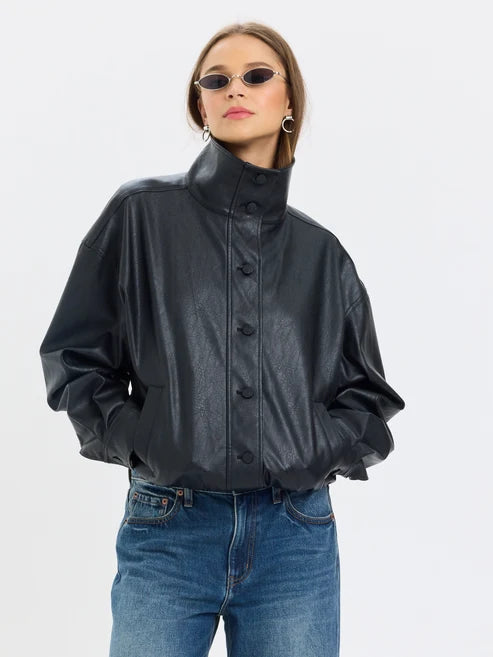 A woman wearing a black faux-leather high-neck jacket with front buttons and voluminous sleeves, paired with blue jeans, against a light gray studio background.
